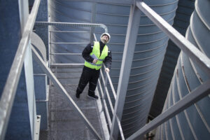 Factory Worker Standing On Metal Platform Between Industrial Storage Tanks And Looking Up For Visual Inspection Of Silos Food Production.