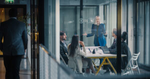 Businesswoman Leads A Strategic Planning Meeting In A Glass Conference Room, Explaining Goals, Reviewing Performance Metrics, Coordinating Tasks, And Encouraging Collaboration As Colleagues Listen