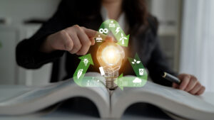 Spark Of Innovation: A Business Person Holding A Light Bulb Over An Open Book With Eco Friendly Icons Representing Sustainability And Energy Solutions Quark
