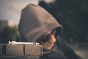Intoxicated Old Man In Hooded Shirt Taking Drugs In City Park On Bench
