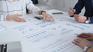 Business Team In A Conference Room Reviewing Financial Reports, Charts And Graphs Across The Table, Collaborating On Strategy, Investment Planning And Growth Initiatives