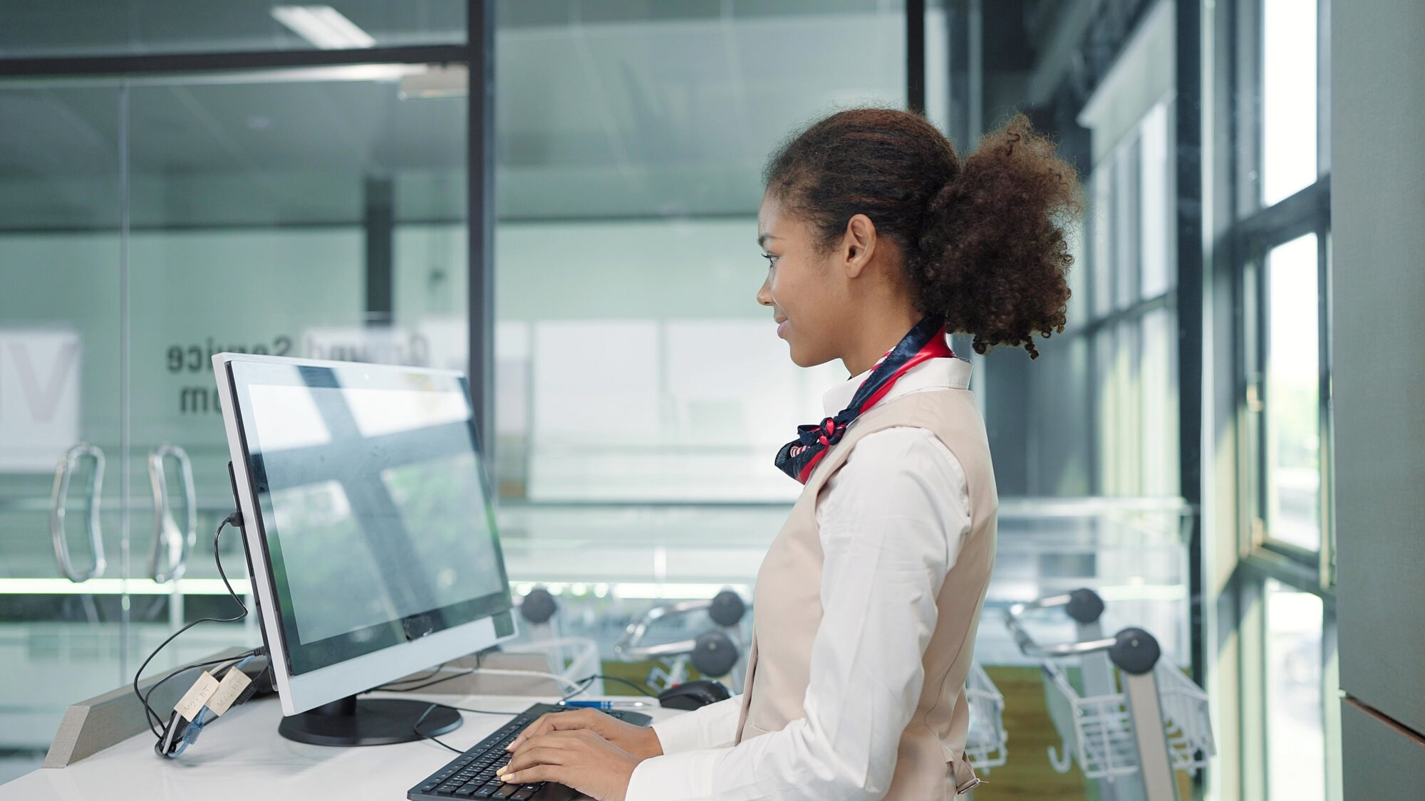 Young American African Women Airline Ground Staff Worker In Uniform Working With Computer In Airport Check In Counter. Ground Hostess Airport Reception At Airline Check In Counter