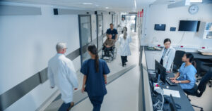 High Angle CCTV Shot With A Busy Hallway With Reception Desk Of A Hospital Building, Diverse Female And Male Staff Members Walking, Working. Professional Nurses, Doctors And Personnel At Work